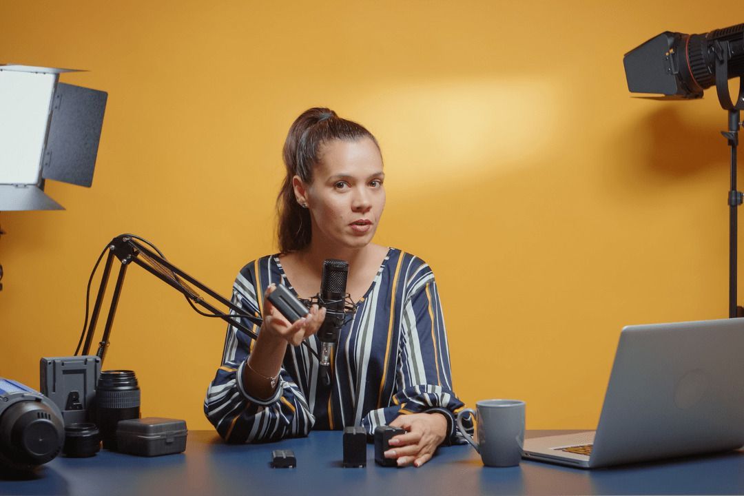 Frau mit Pferdeschwanz und gestreifter Bluse sitzt am Tisch und hat ein Podcast-Mikrofon Gelenkarmstativ vor sich. Sie schaut den Betrachter an. Auf dem Tisch stehen Laptop, Tasse sowie Kamerautensilien und im Hintergrund zwei Lichtquellen.