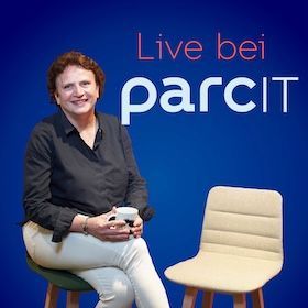 Eine Frau sitzt mit Kaffeetasse in den Händen auf einem Stuhl, hat die Beine übereinandergeschlagen und lächelt zum Betrachter. Daneben ein leerer Stuhl. Schriftzug: Live bei parcIT. Podcast erstellen.
