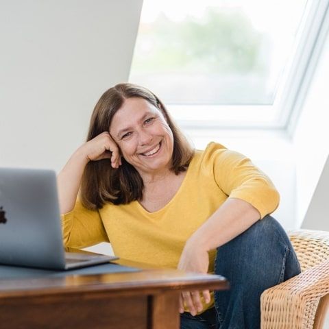 Podcasterin am Tisch mit Notebook stützt ihren Kopf seitlich auf die Hand und lächelt. Fenster im Hintergrund.