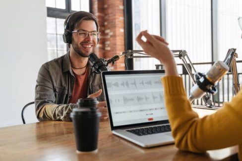 Lächelnder Podcaster am Podcast -Mikrofon, der seinem Podcast-Host gegenüber sitzt. Beide Personen nehmen gerade eine Podcast-Episode mit ihrem Aufnahmeprogramm auf.