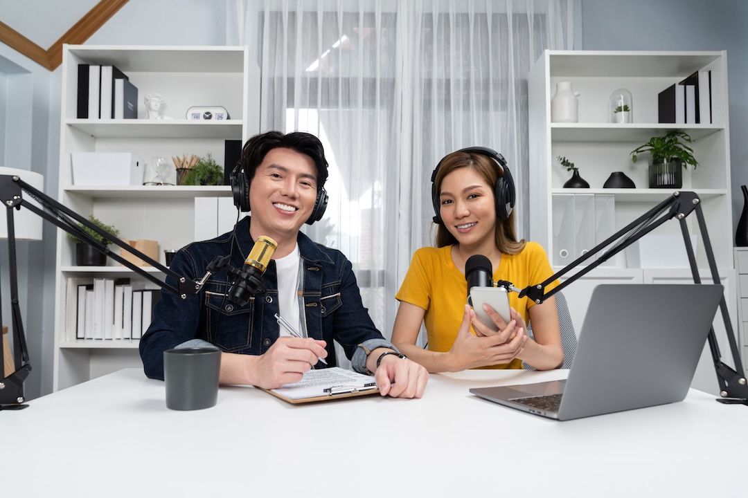 Ein Mann und eine Frau sitzen an ihren Podcast-Mikrofonen und tragen Podcast-Kopfhörer. Sie machen eine Podcast-Aufnahme. Auf dem Tisch steht ein Laptop und eine Kaffeetasse. Sie hält ein Smartphone in der Hand und er schreibt das Podcast-Skript. Im Hintergrund zwei Regale mit Büromaterialien.